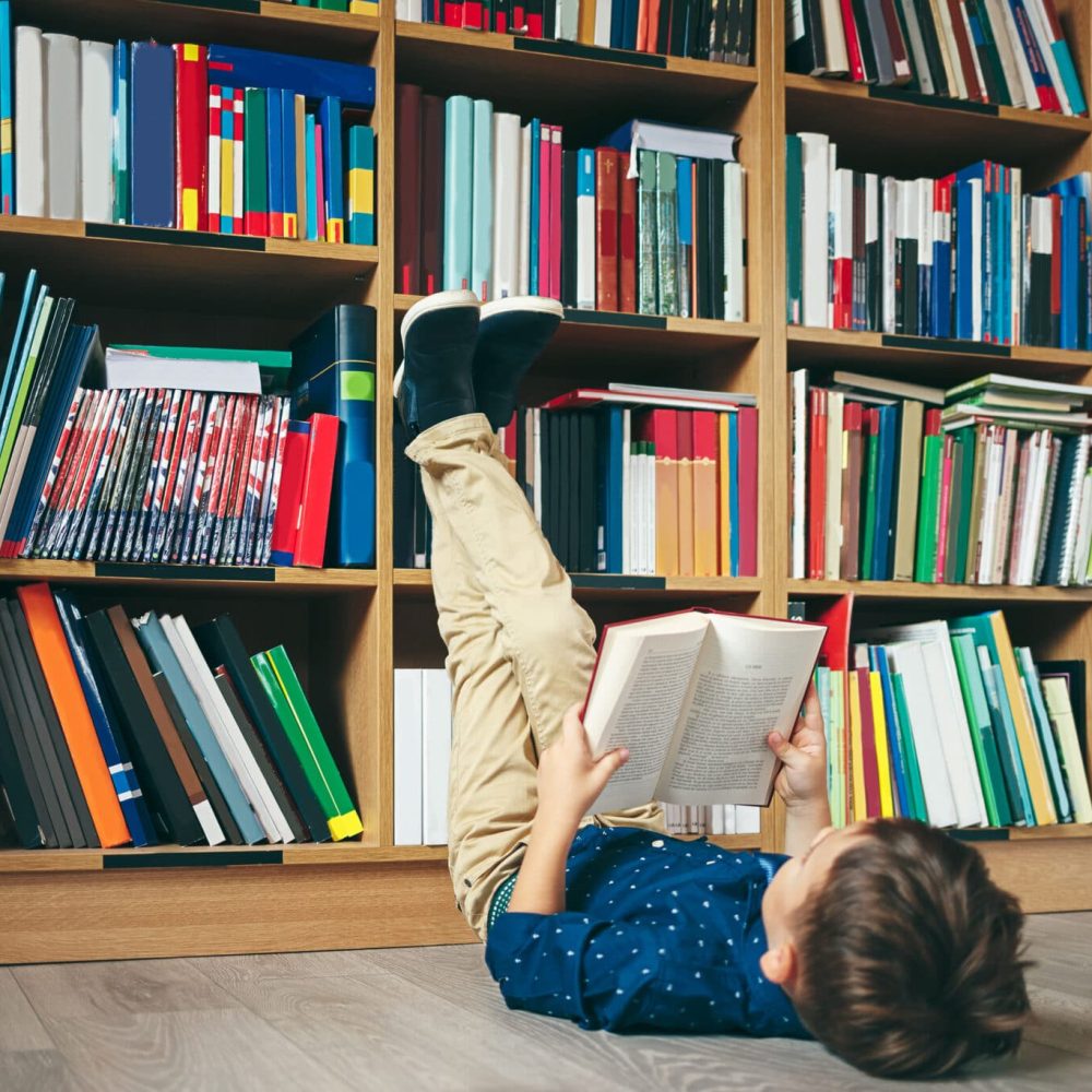 Boy laying on  the floor with the feet up, reading a book against multi colored bookshelf in library. Education, Knowledge, Bookstore, Lecture. Pupil holds a book in his hands.